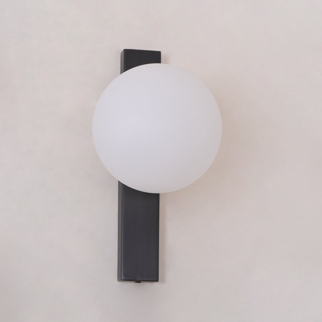 Frosted white globe on a flat black bar, this Clérachic wall light is shown unlit and close to the frame against a pale wall.
