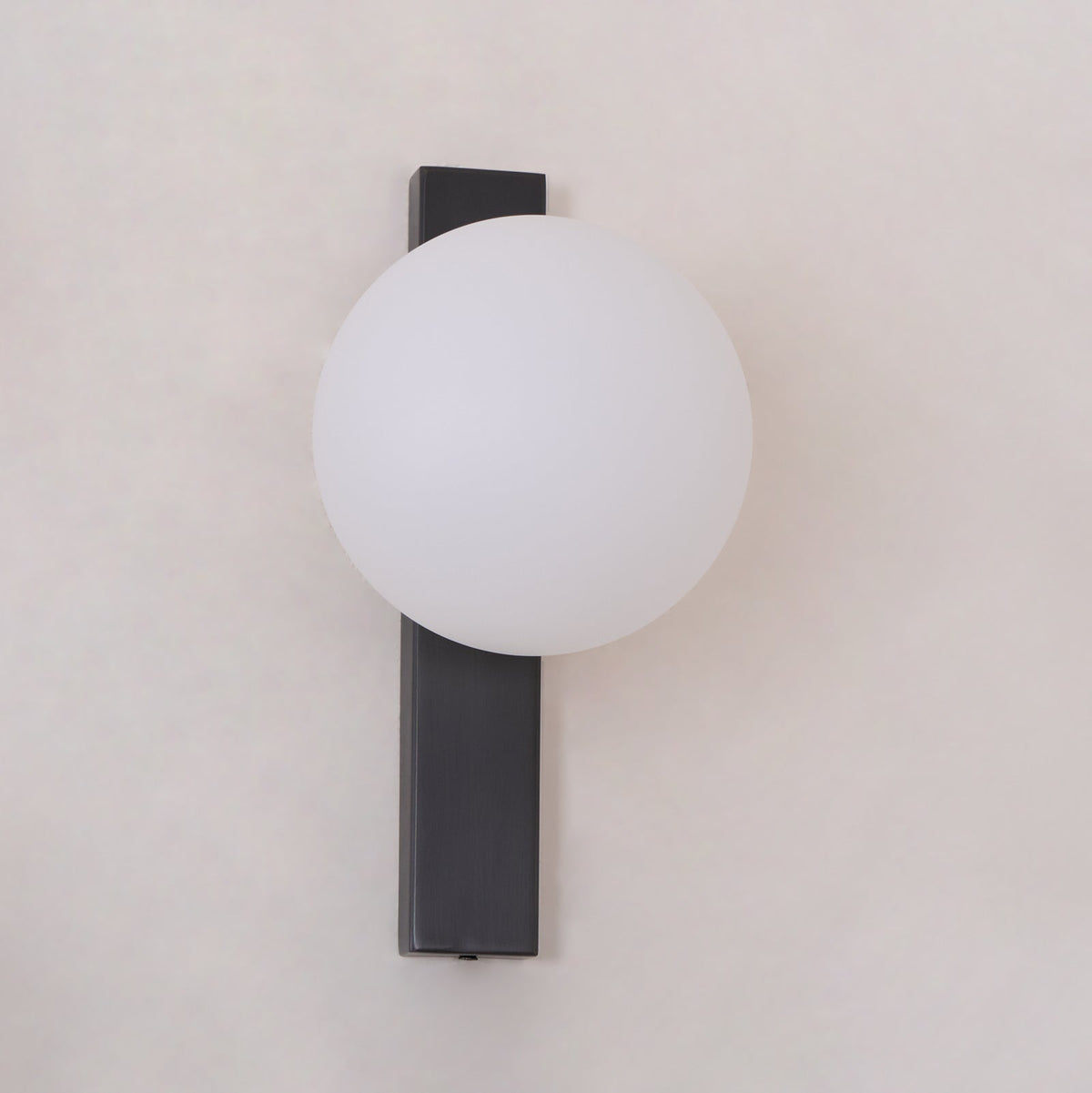 Frosted white globe on a flat black bar, this Clérachic wall light is shown unlit and close to the frame against a pale wall.