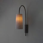 Lit Clérachic wall light with a curved black iron arm and a grey fabric shade on a dark grey wall, glowing softly.