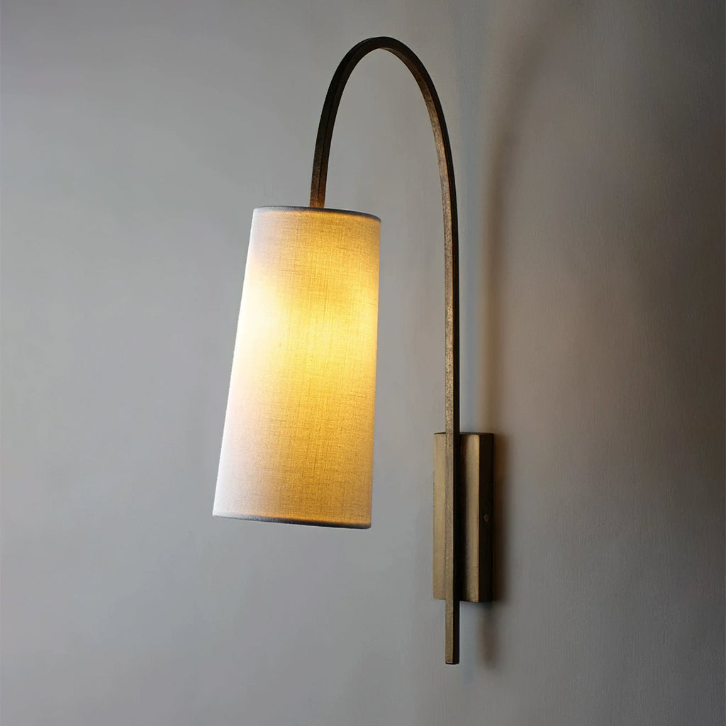 This Clérachic wall light lit, its white fabric shade glowing warm on a gold-toned arm that curves over from a slim backplate.