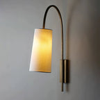 This Clérachic wall light lit, its white fabric shade glowing warm on a gold-toned arm that curves over from a slim backplate.