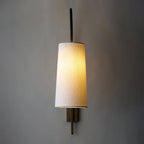 A tapered white fabric shade lit warm on this Clérachic wall light, hanging from a bronze-toned arm against a deep grey wall.