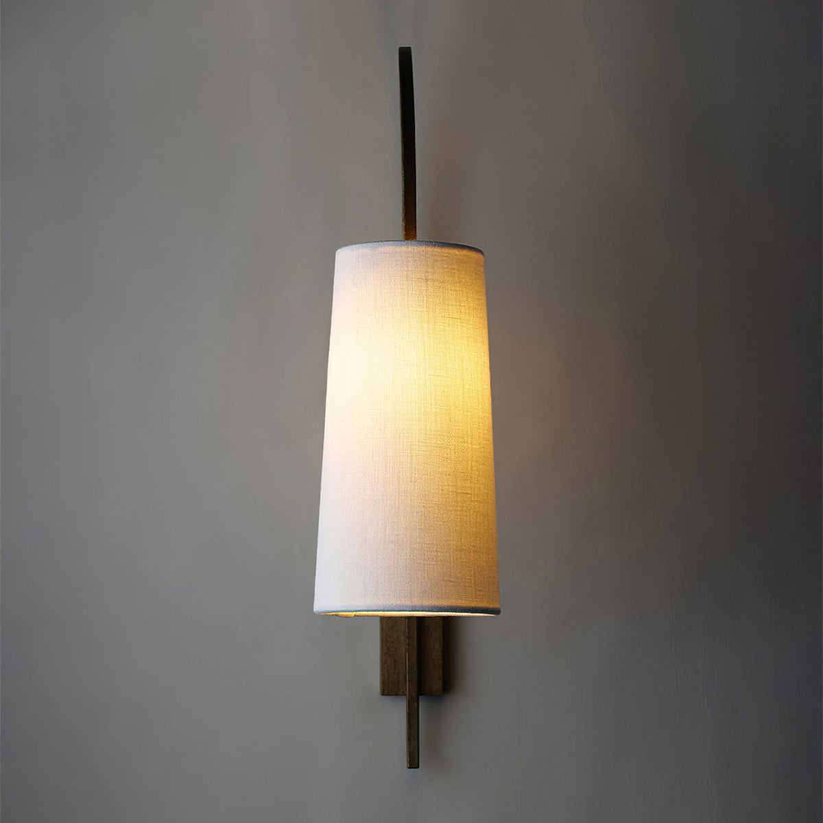 A tapered white fabric shade lit warm on this Clérachic wall light, hanging from a bronze-toned arm against a deep grey wall.
