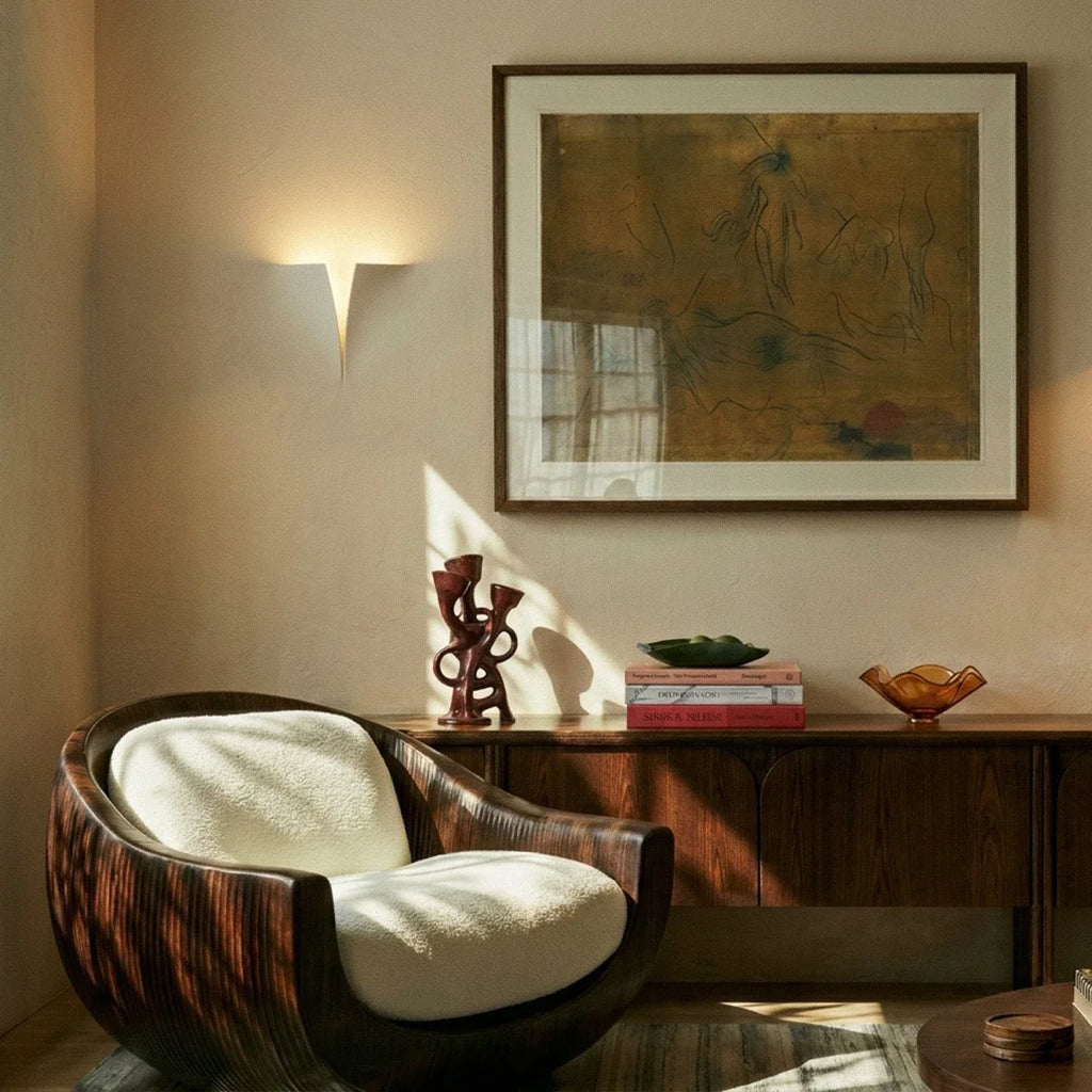 Clérachic white wall light washing warm light up a cream wall above a wooden sideboard and a rounded wooden armchair.