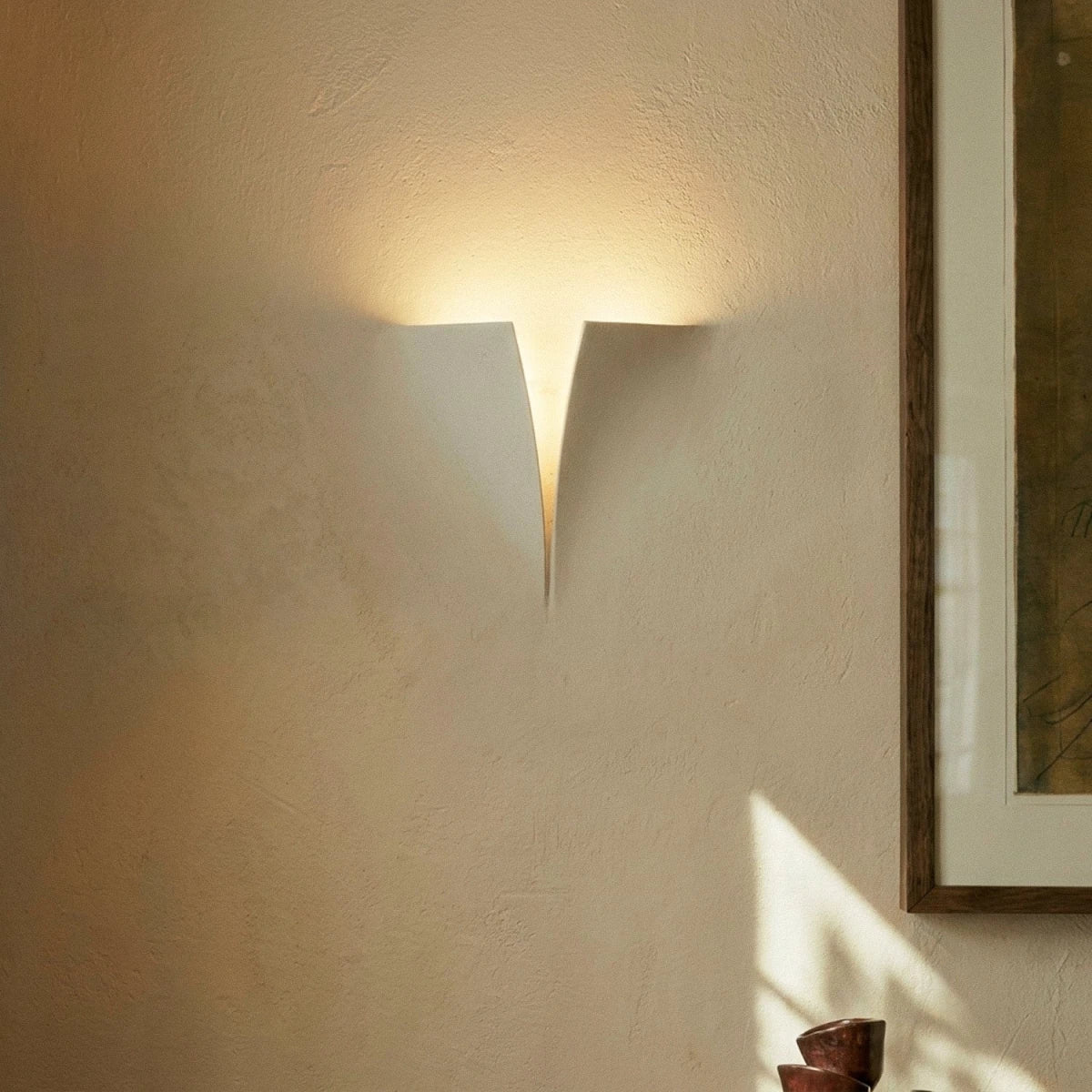 Close view of a Clérachic white wall light casting a warm upward glow over a textured plaster wall beside a framed artwork.