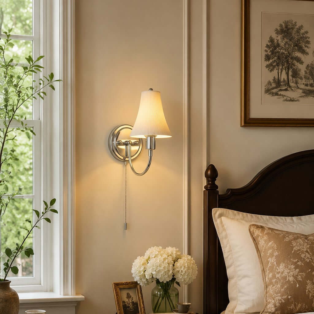 Lit fluted-shade wall light beside a sash window and a dark wooden bedhead, glowing warm over cream bedding and white hydrangeas, from Clérachic.