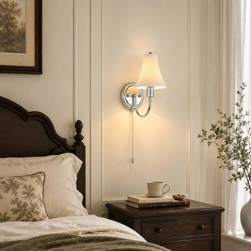 Chrome-armed wall light with a fluted shade above a dark bedside chest, a stacked pair of books and a cup, in a panelled bedroom from Clérachic.