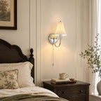 Chrome-armed wall light with a fluted shade above a dark bedside chest, a stacked pair of books and a cup, in a panelled bedroom from Clérachic.