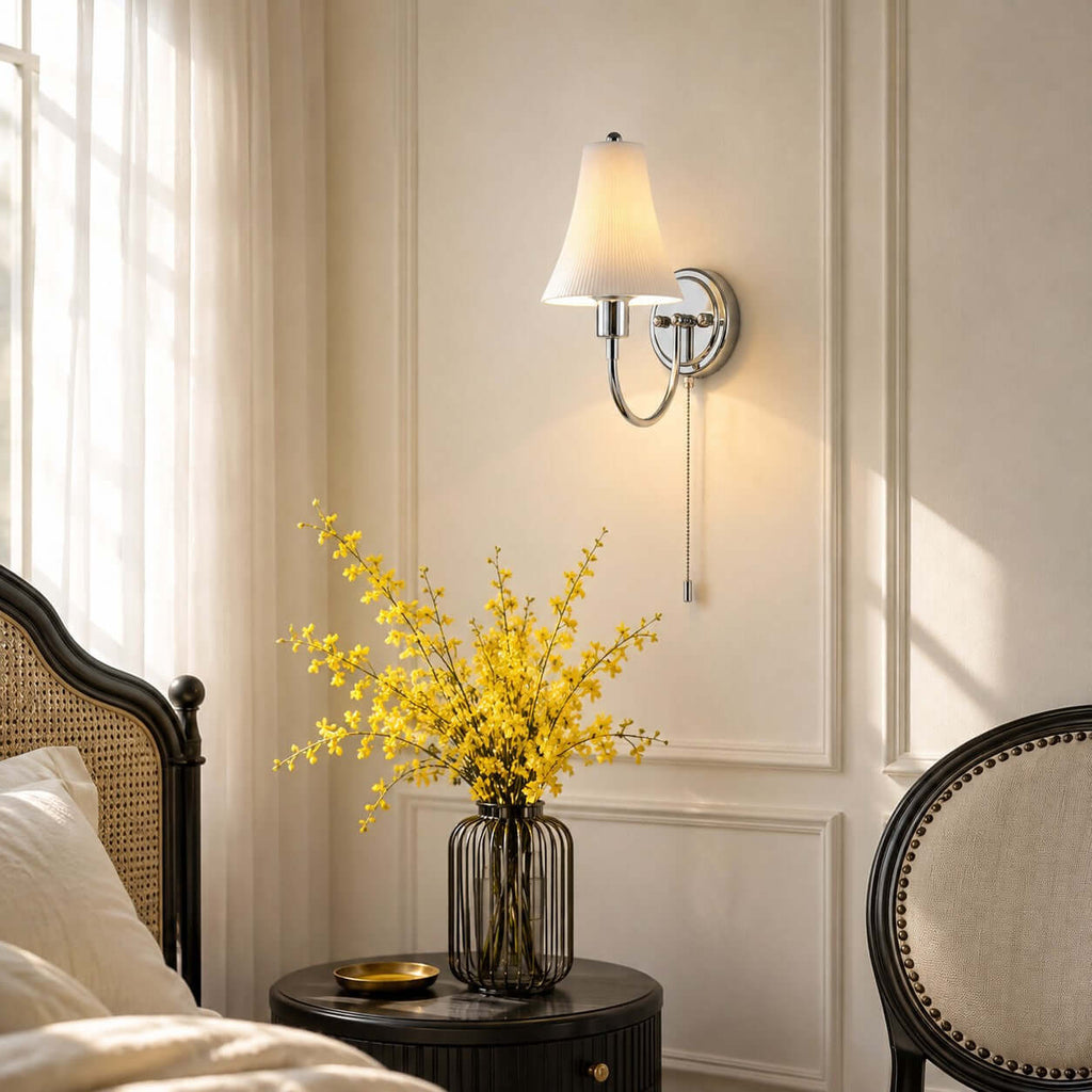 Seen from the side, this Clérachic sconce lights a panelled wall beside a round dark table holding a vase of yellow blossom.