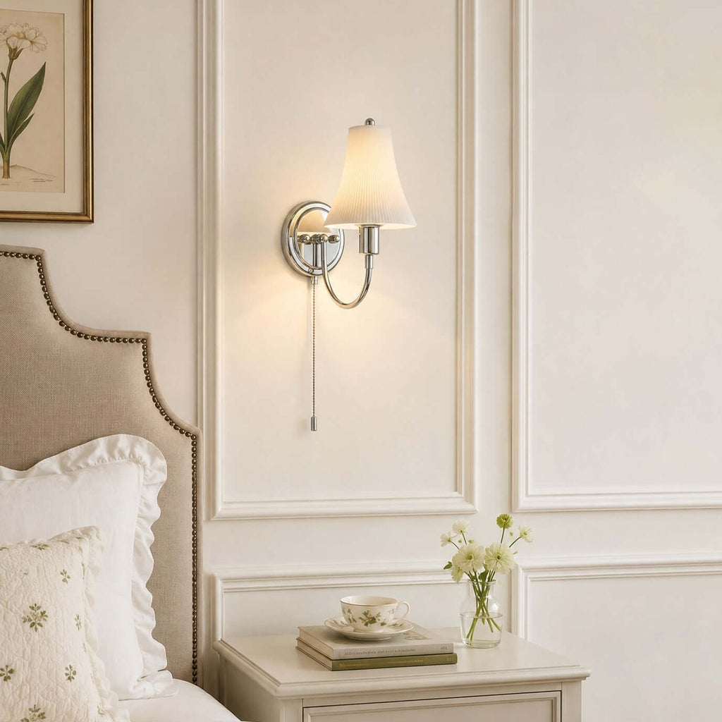 A Clérachic wall sconce on white panelling above a studded headboard, with a ruffled pillow and a small posy on the table beside it.
