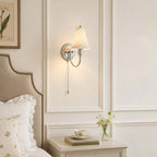 A Clérachic wall sconce on white panelling above a studded headboard, with a ruffled pillow and a small posy on the table beside it.