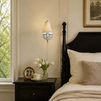 This Clérachic sconce, lit, with a polished chrome backplate, hangs over a black four-poster bed and a nightstand with white tulips.