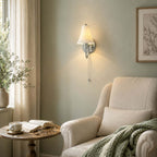 On a soft sage-green wall above a reading corner, this Clérachic light shines beside a cream armchair, a green knit throw and an open book.