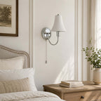 Daylight falls across a white wall where this Clérachic chrome-armed sconce hangs above a pale oak bedside chest, stacked books and a stone vase.
