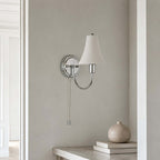Unlit ceramic shade and polished chrome arm of this Clérachic wall light against a grey plaster wall, with a stone vase and books on a ledge below.