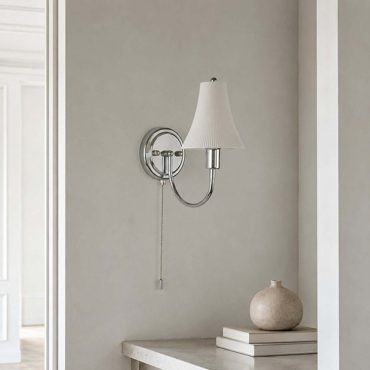 Unlit ceramic shade and polished chrome arm of this Clérachic wall light against a grey plaster wall, with a stone vase and books on a ledge below.