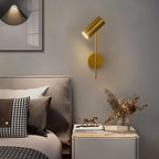 Clérachic gold wall reading light over a pale bedside cabinet with books, dried stems and a grey upholstered headboard.