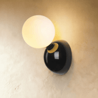 Clérachic black wall lamp with a lit frosted globe on a gloss black dome plate with a brass collar, on plaster.