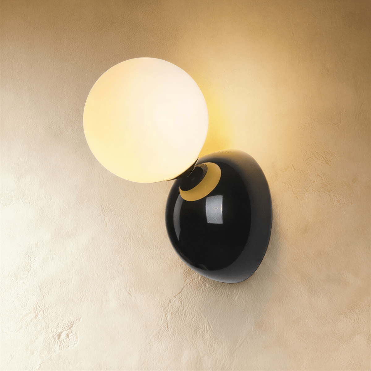 Clérachic black wall lamp with a lit frosted globe on a gloss black dome plate with a brass collar, on plaster.