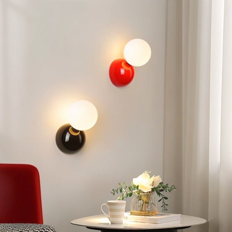 Clérachic red and black wall lamps with lit frosted globes on a pale wall above a side table with flowers.