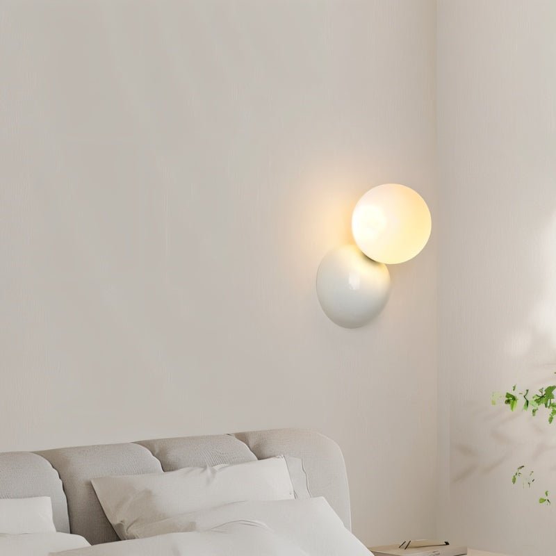 Clérachic white wall lamp with a lit frosted globe on a cream wall above a grey upholstered bed and white pillows.