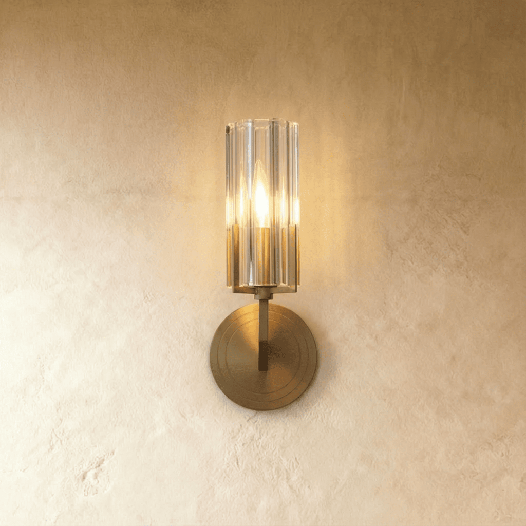 Clérachic single-shade wall light with a fluted glass cylinder and brass round backplate, glowing warmly on a limewashed wall.