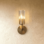 Clérachic single-shade wall light with a fluted glass cylinder and brass round backplate, glowing warmly on a limewashed wall.