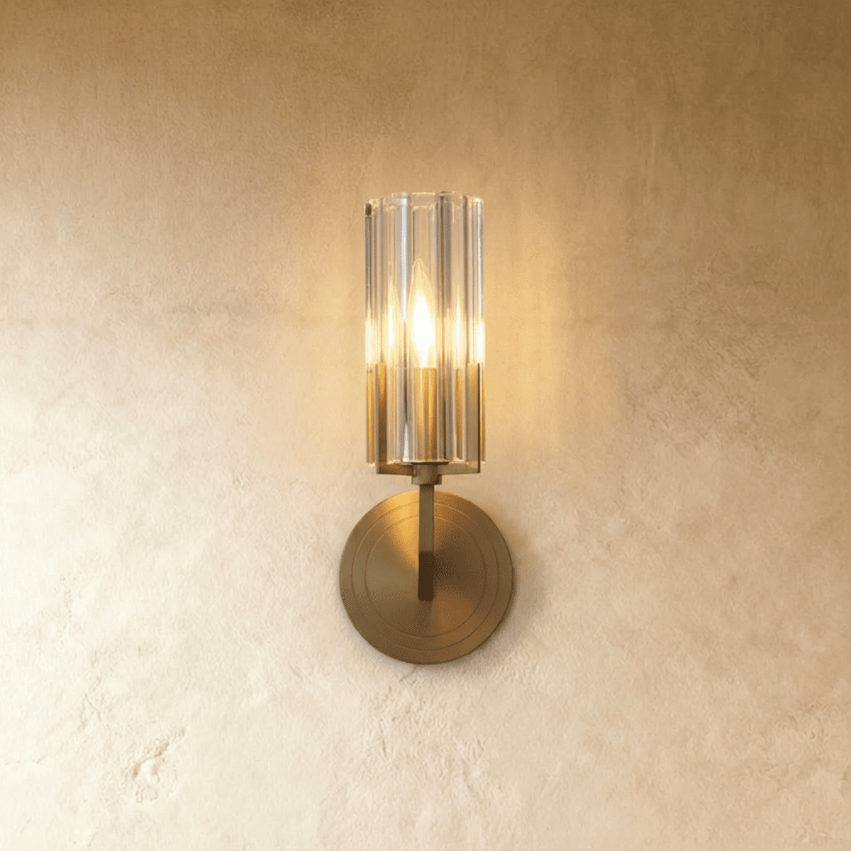 Clérachic single-shade wall light with a fluted glass cylinder and brass round backplate, glowing warmly on a limewashed wall.