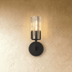 Clérachic black wall light with a fluted glass cylinder shade lit warmly, mounted on a round stepped backplate against plaster.