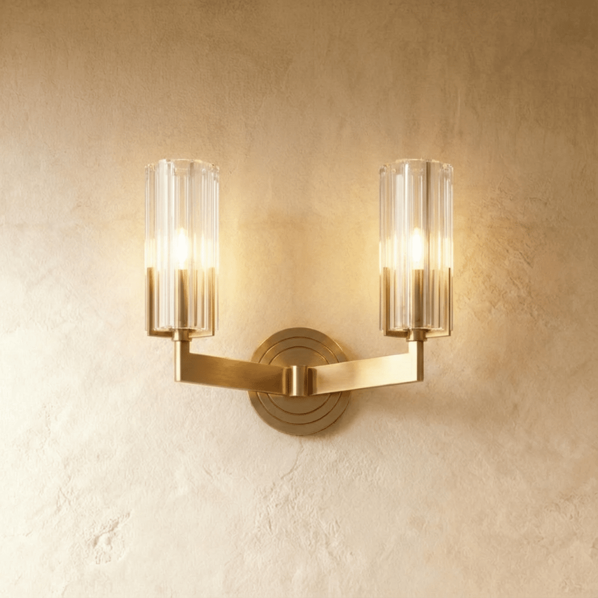 Clérachic twin-arm brass wall light with two fluted glass shades lit warmly, mounted on a limewashed wall.