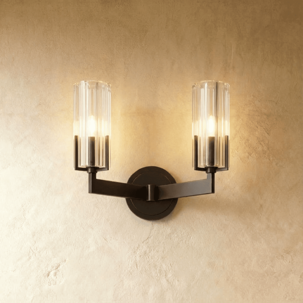 Clérachic twin-arm black wall light with two lit fluted glass shades on angular arms, set against a warm plaster wall.