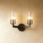 Clérachic twin-arm black wall light with two lit fluted glass shades on angular arms, set against a warm plaster wall.