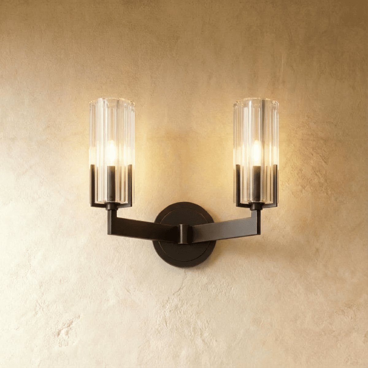 Clérachic twin-arm black wall light with two lit fluted glass shades on angular arms, set against a warm plaster wall.