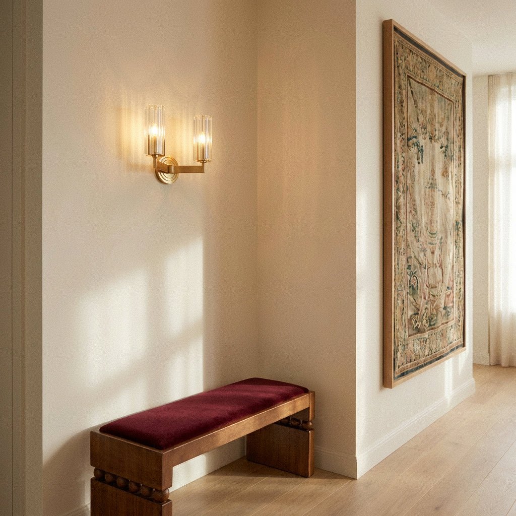 Clérachic twin-arm brass wall light glowing in a bright hallway beside a tapestry, above a walnut bench with a burgundy cushion.
