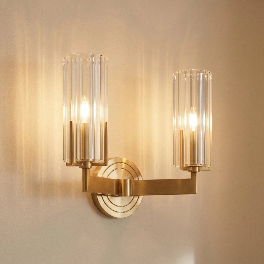 Close view of the Clérachic twin-arm brass wall light, two lit candle bulbs inside fluted glass shades on a stepped round backplate.