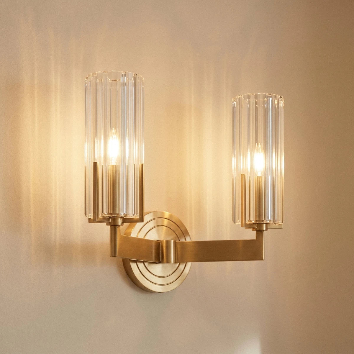 Close view of the Clérachic twin-arm brass wall light, two lit candle bulbs inside fluted glass shades on a stepped round backplate.