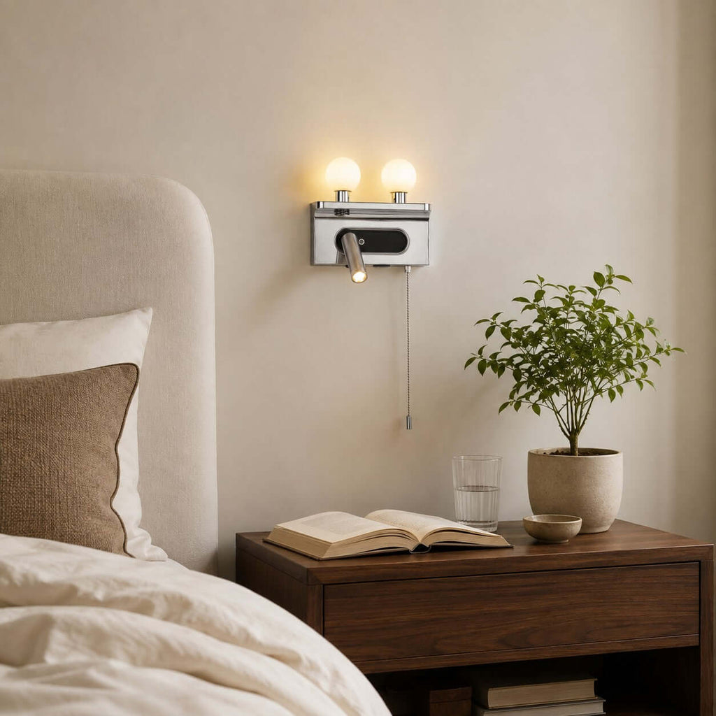 A chrome Clérachic wall light with a pull chain sits over a walnut bedside table with an open book and a small plant.