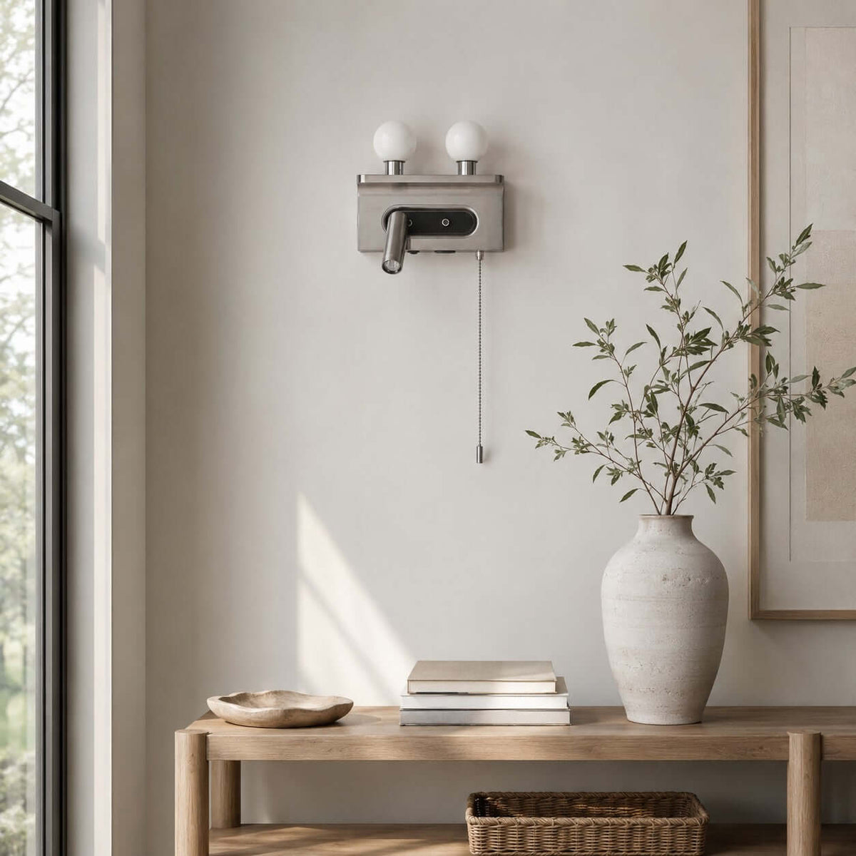 A nickel Clérachic wall light with two unlit globes hangs by a tall window above a wooden bench and a stack of books.