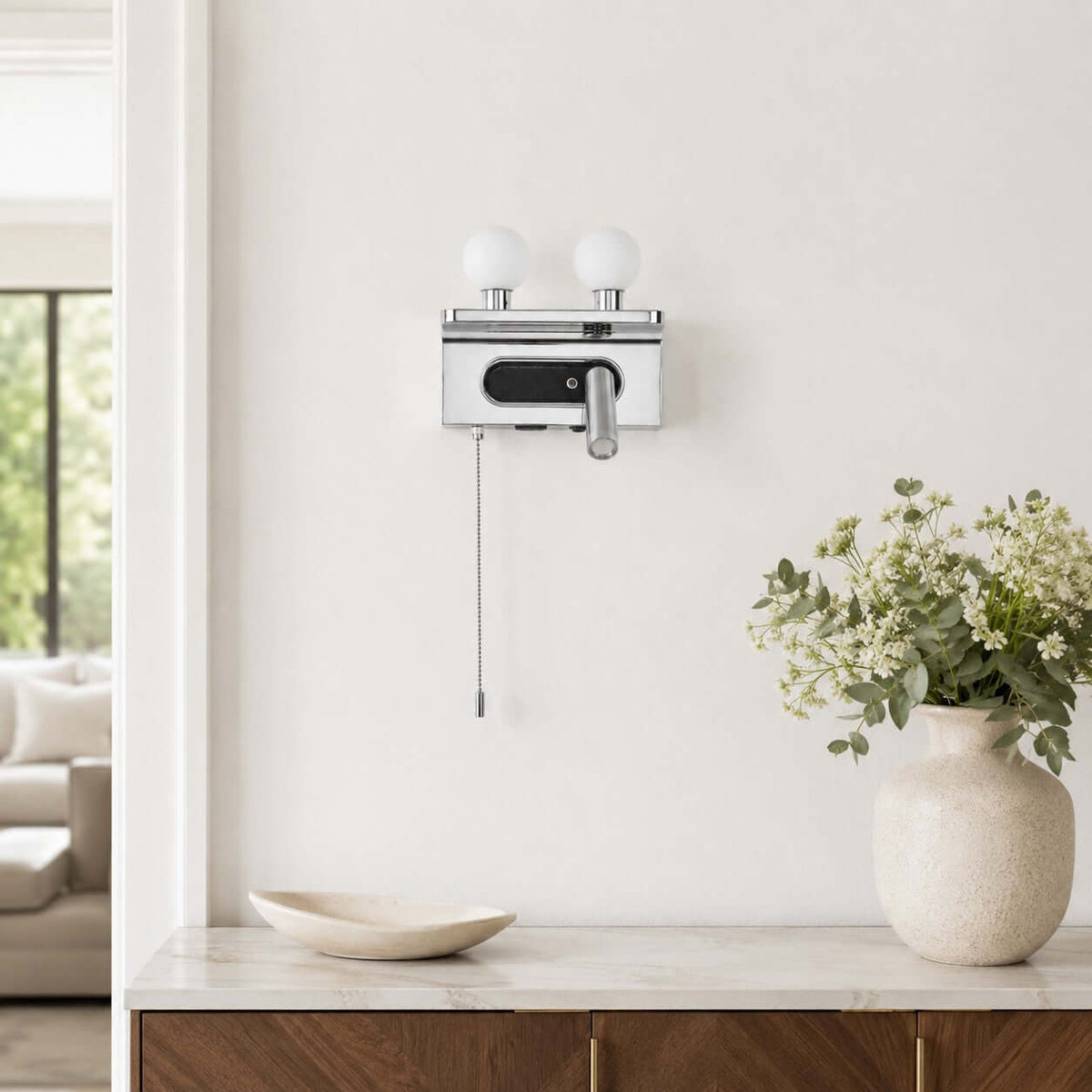 A chrome Clérachic wall light with unlit white globes hangs above a marble-topped sideboard with a vase of flowers.