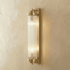 Clérachic wall light in brushed gold with a ribbed glass rod cylinder and two lit bulbs, on a limewashed wall.