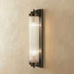 Clérachic wall light in dark bronze-black with a ribbed glass rod cylinder and two lit bulbs, on limewash.
