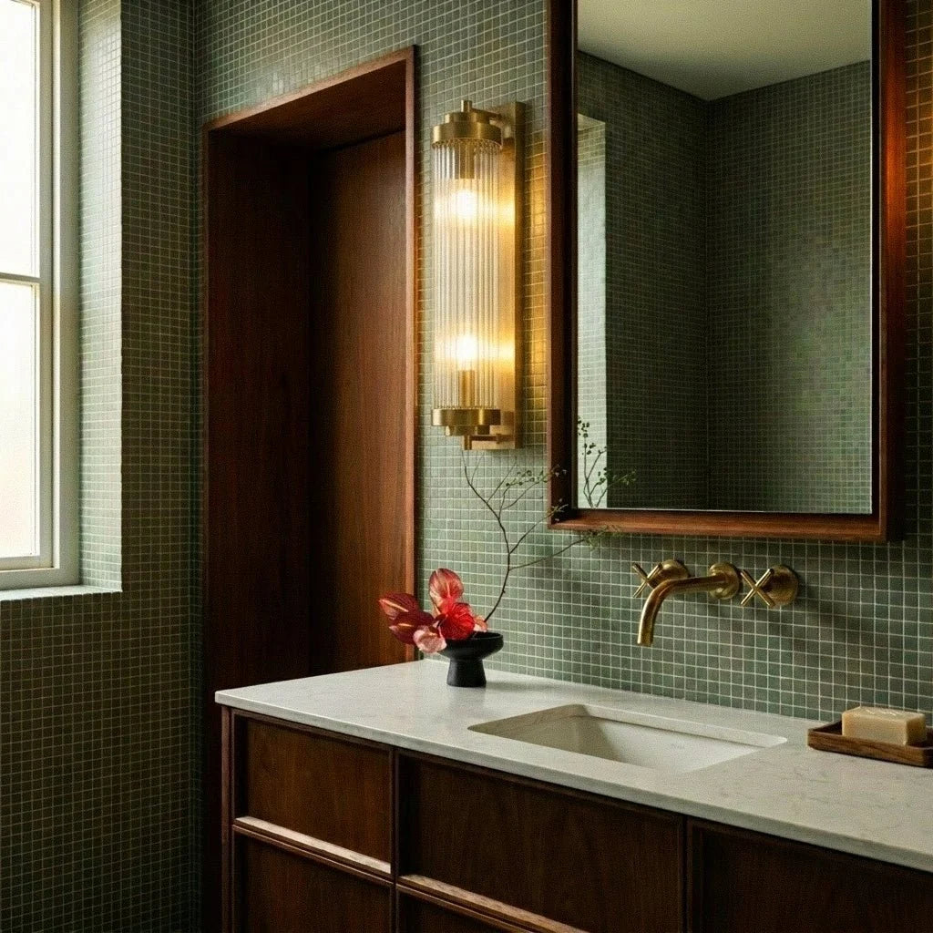 Clérachic gold wall light with ribbed glass rods beside a framed mirror over a walnut vanity and green mosaic tiles.
