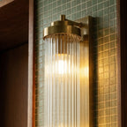 Close view of the Clérachic gold wall light's brass cap, ribbed glass rods and lit bulb against green mosaic tile.