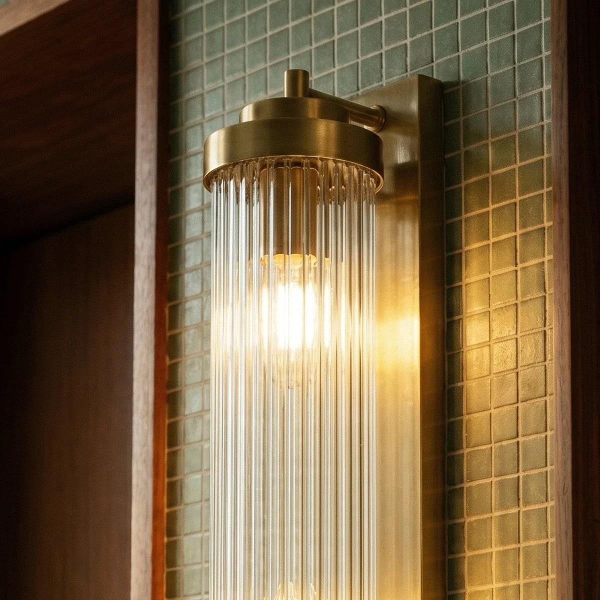Close view of the Clérachic gold wall light's brass cap, ribbed glass rods and lit bulb against green mosaic tile.