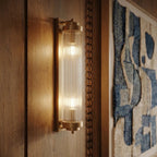 Clérachic gold wall light with two lit bulbs behind ribbed glass rods, mounted on warm oak wall panelling.