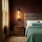 Clérachic dark bronze-black wall light with ribbed glass rods glowing beside a walnut bedside table and striped headboard.