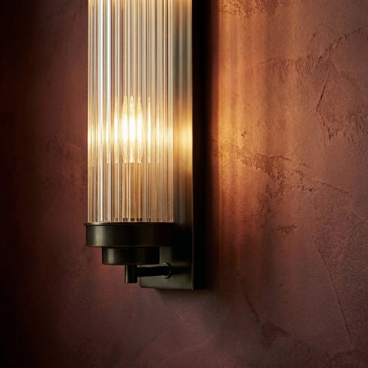 Close view of the Clérachic dark bronze-black wall light's base, ribbed glass rods and glowing bulb on a plum wall.