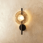 Clérachic wall light with a single amber glass disc and frosted bulb glowing on a slim black rod with a round backplate.