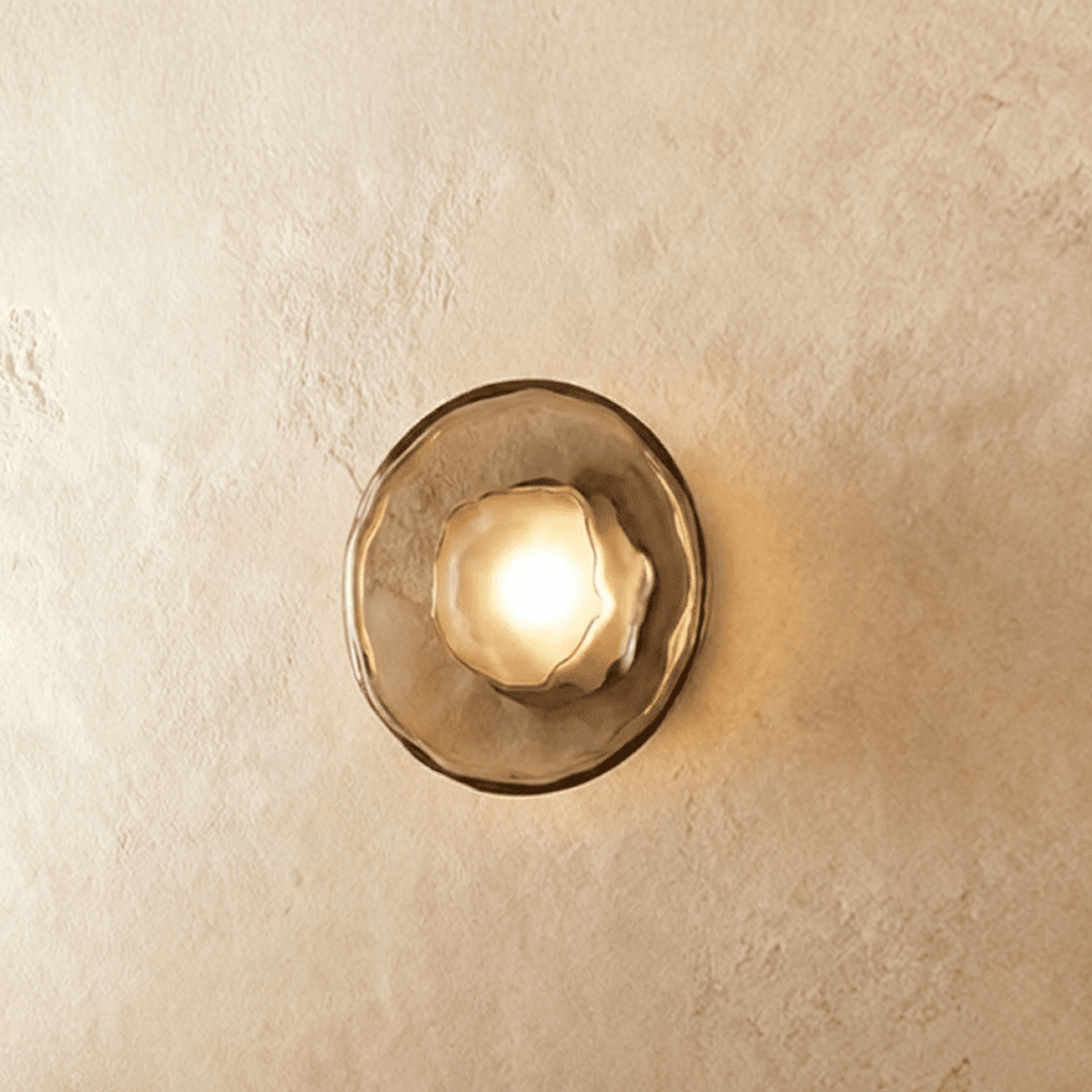 Clérachic round wall light with a smoky glass disc and frosted glass bulb cover, glowing warm on a plaster wall.