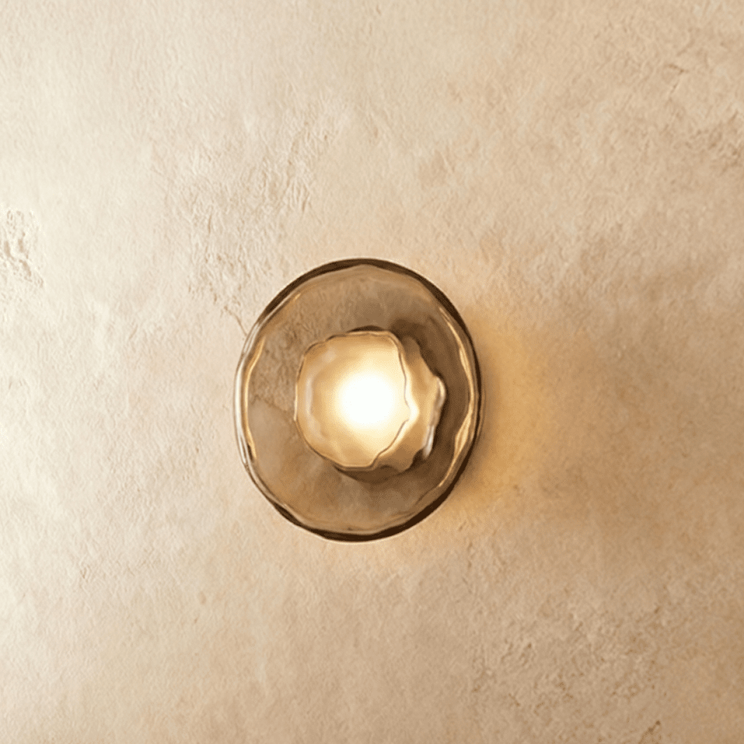 Clérachic round wall light with a smoky glass disc and frosted glass bulb cover, glowing warm on a plaster wall.